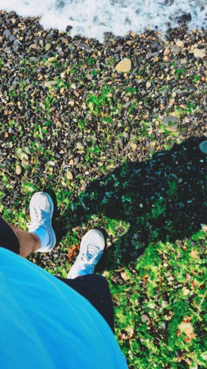 Looking down at white sneakers on a pebble beach with green moss, the edge of a wave at the top