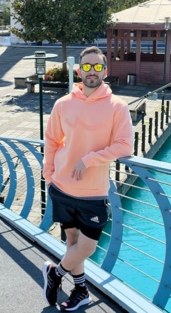 Amir in a peach hoodie, shorts and yellow mirrored sunglasses, leaning on a blue railing above turquoise water