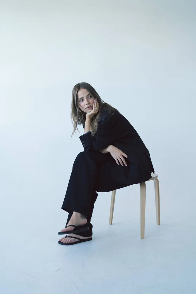 A woman in a black suit sitting on a wooden stool against a pale studio wall, chin in her hand, looking over her shoulder