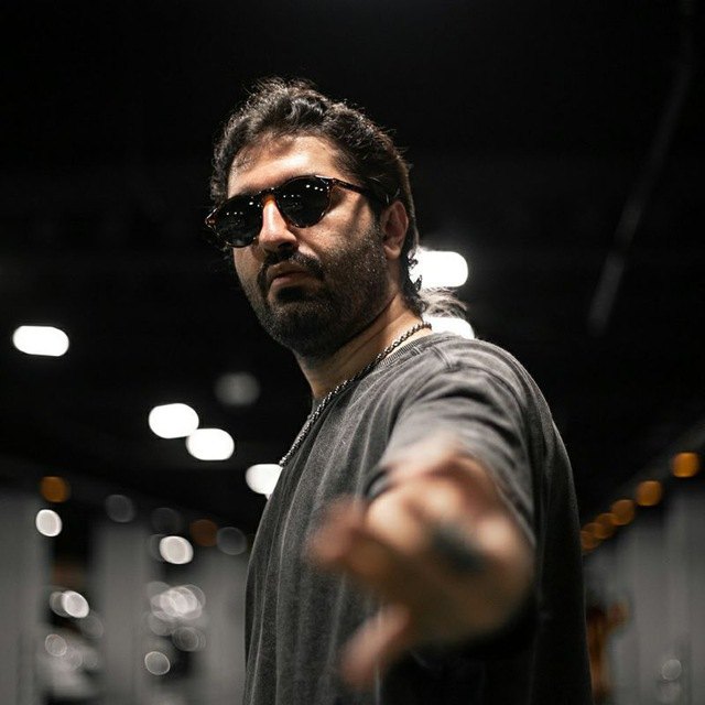 Farzam in round sunglasses and a dark T-shirt, reaching a hand toward the camera in a softly lit hall