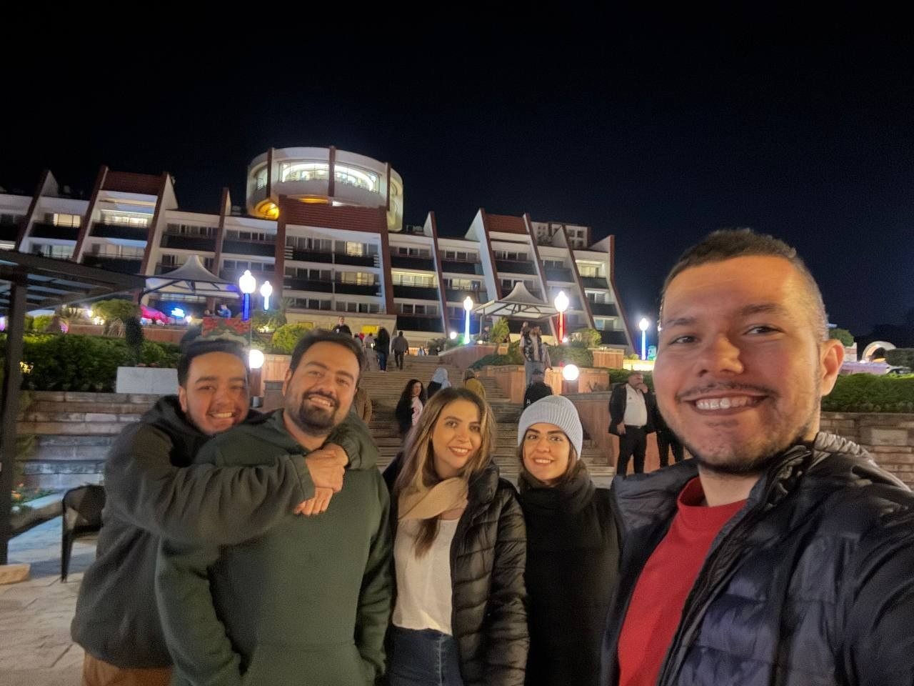 A night-time selfie of five friends in front of a lit-up hotel