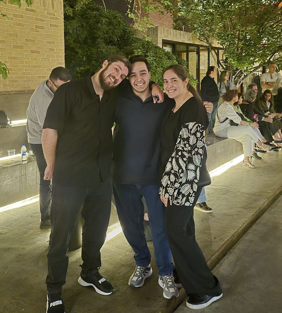 Three friends with their arms around each other on a lit walkway under trees