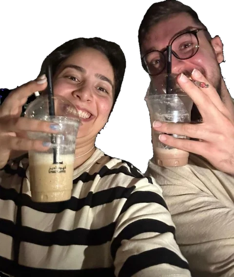 Two friends laughing at night, each holding up an iced coffee with a straw