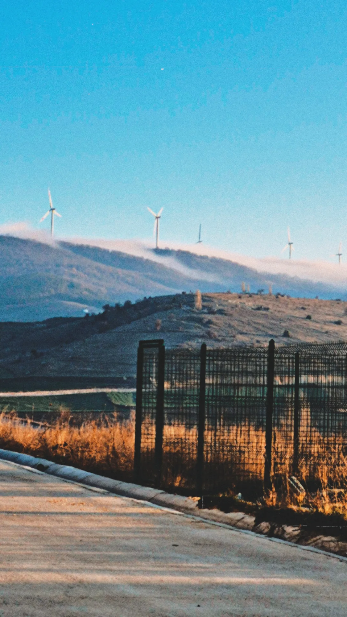 A road beside a fence, with foggy hills and wind turbines under a blue sky