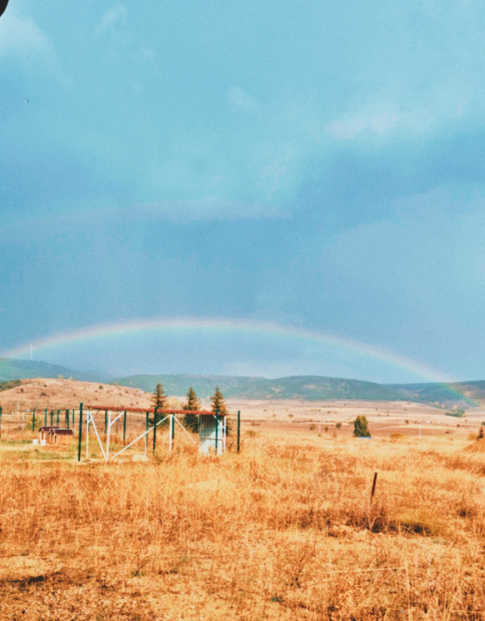 A faint rainbow over a dry golden field with a fence and a few trees