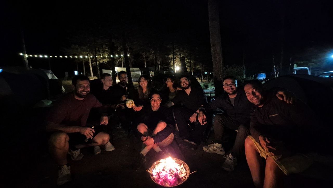 Friends smiling around a small campfire at night, with string lights and tents behind them