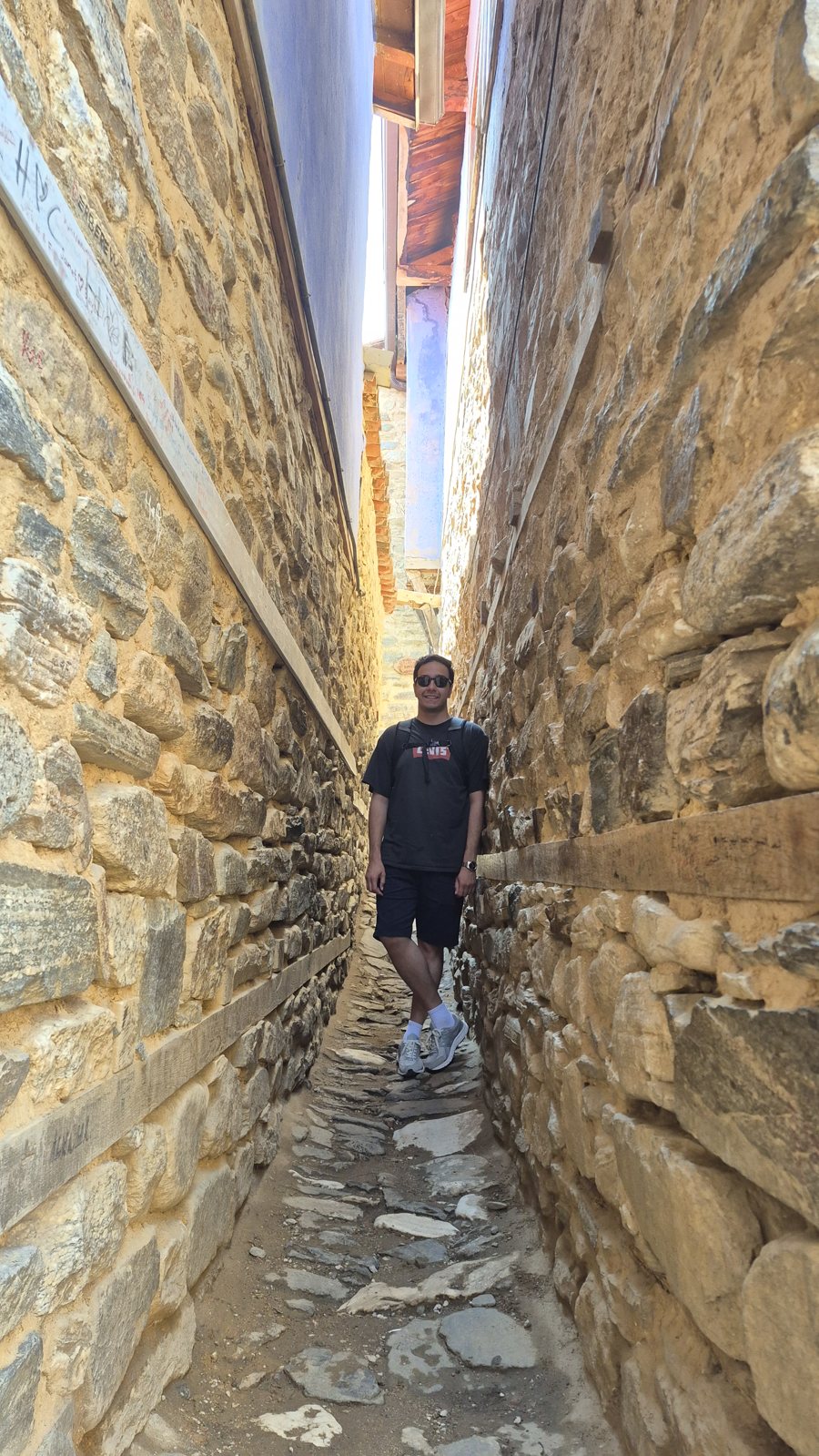 Sina in sunglasses, a black T-shirt and shorts, standing in a narrow stone alley under a sliver of blue sky