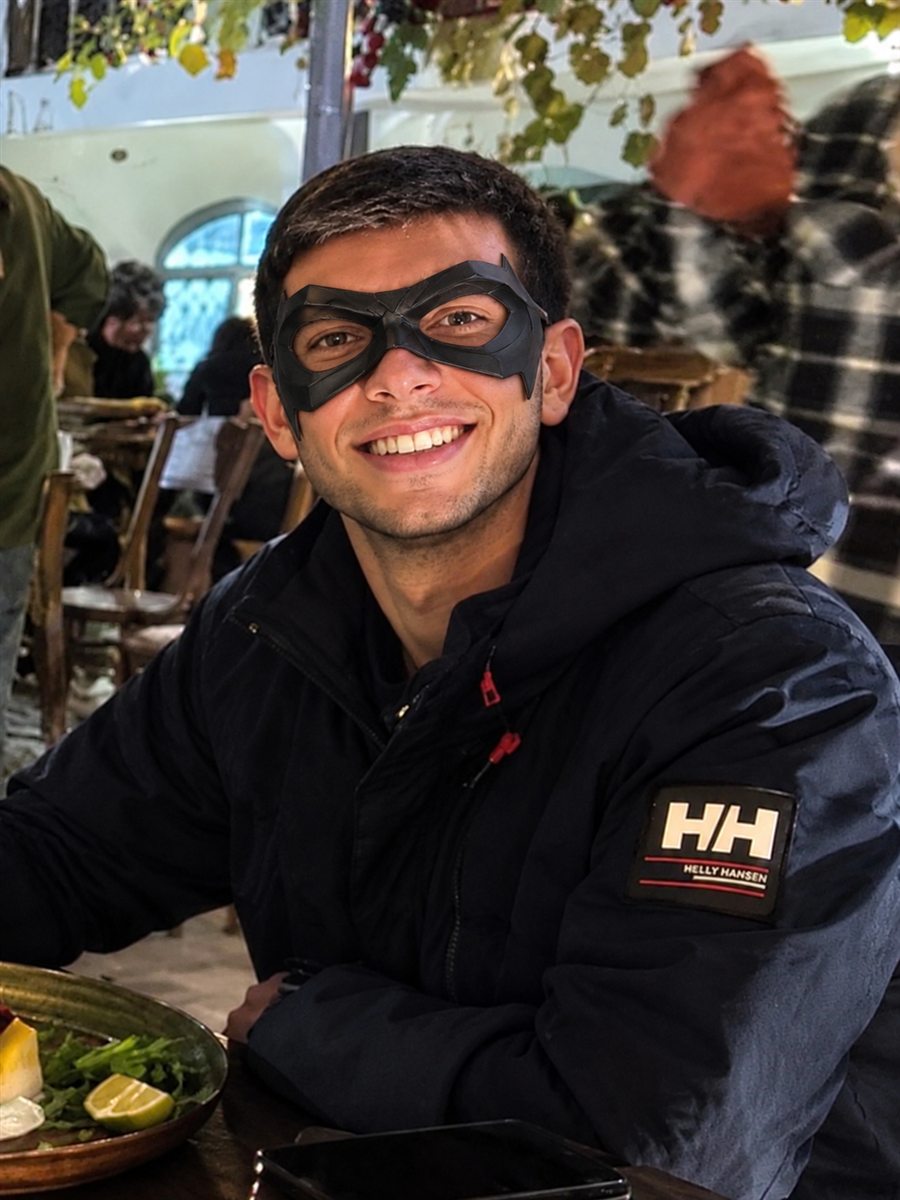 Jorge in a navy jacket and black superhero mask, smiling at a café table