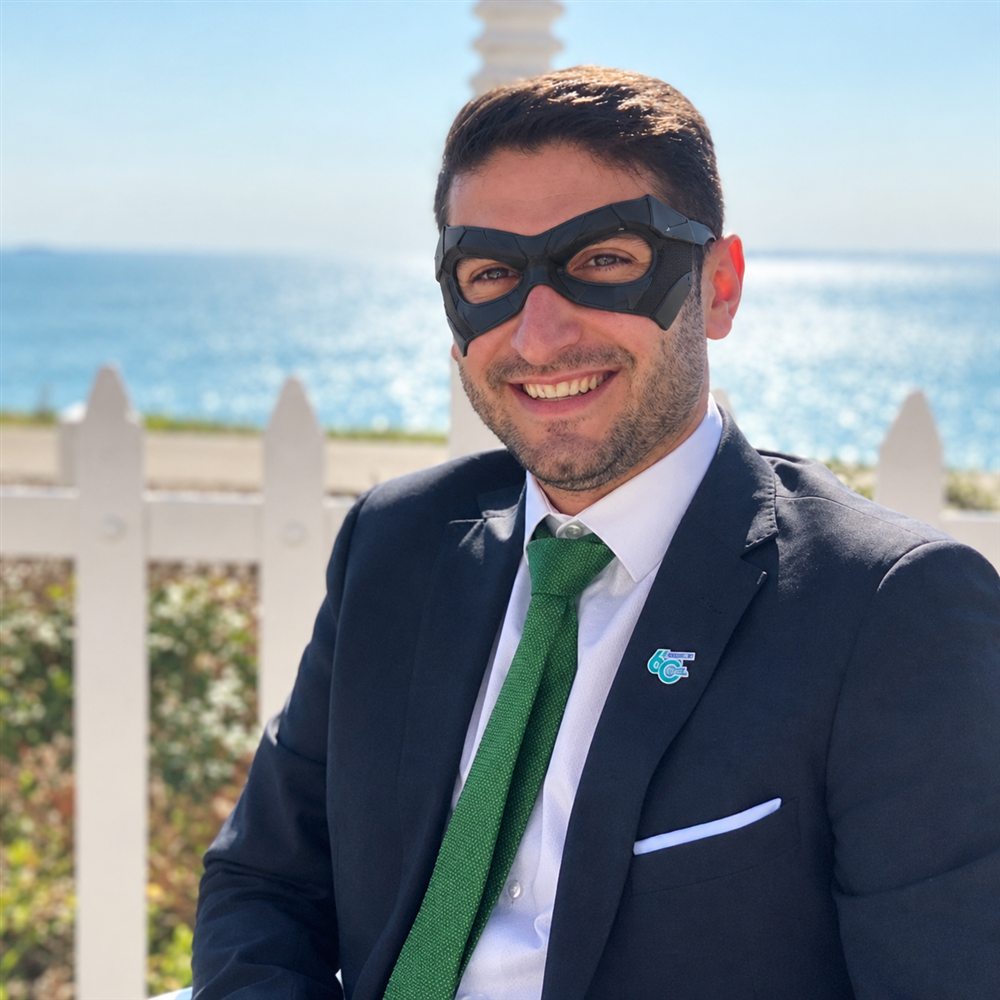 Ruben in a dark suit, green tie and black superhero mask, smiling in front of a white fence and the sea