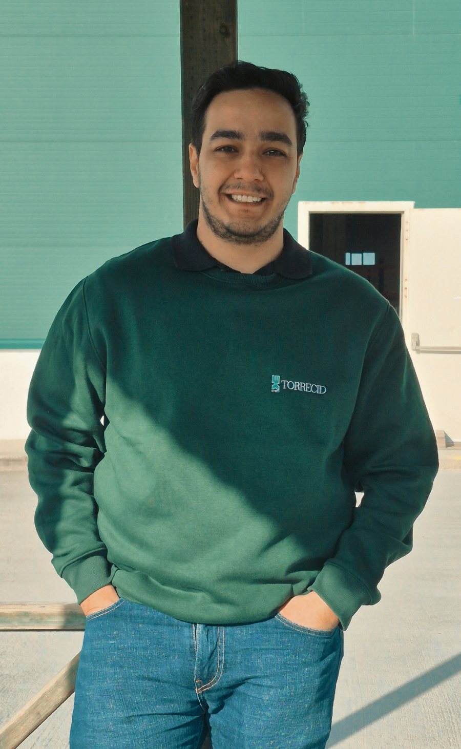 Sina smiling in a green Torrecid sweatshirt and jeans, hands in pockets, in front of a teal warehouse wall